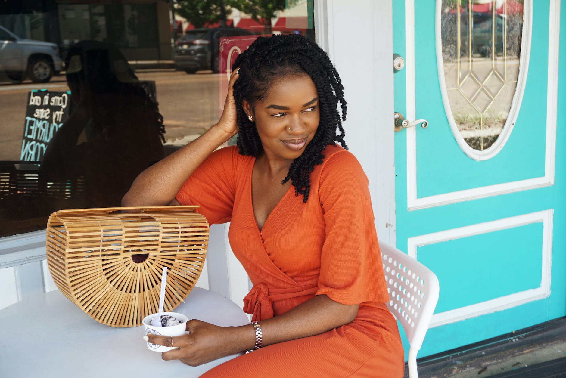 photo of smiling woman in orange v neck short sleeved dress sitting outside store holding ice cream while looking away